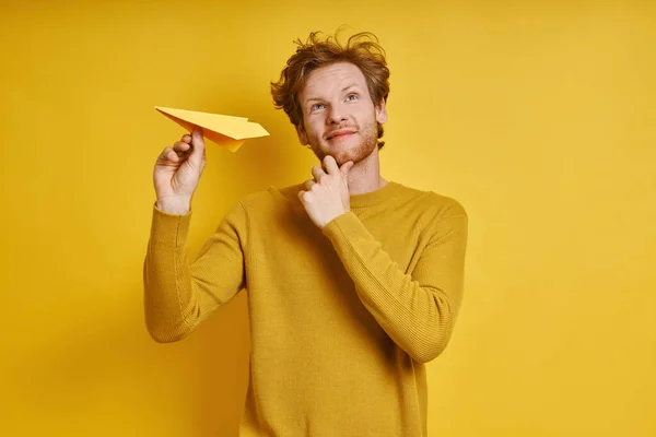 Thoughtful redhead man holding paper airplane while standing against yellow background