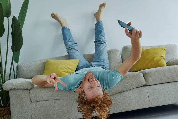 Podekscytowany Rudzielec Trzyma Smartfon Gestykuluje Relaksując Się Kanapie Domu — Zdjęcie stockowe