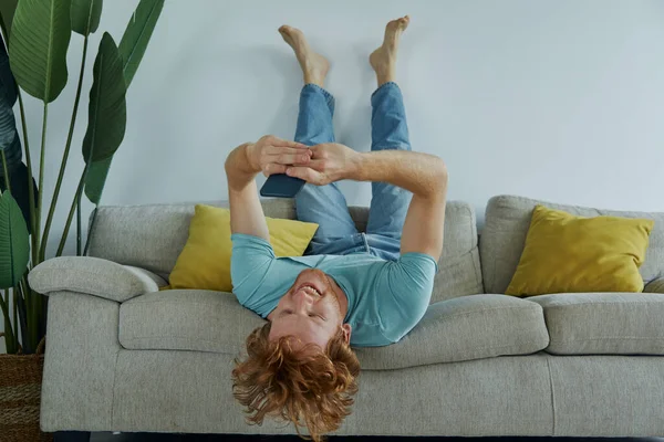 快乐的红头发男人在家里舒服地坐在沙发上 一边用智能手机 — 图库照片