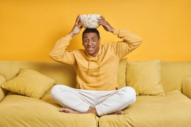 Sarı arka planlı koltukta dinlenirken kafasında patlamış mısır taşıyan oyuncu.