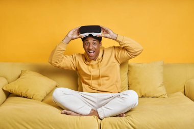 Sarı arka planlı kanepede dinlenirken sanal gerçeklik gözlüğü takan mutlu genç adam.