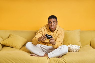 Sarı arka planda kanepede oturmuş video oyunu oynayan konsantre bir adam.