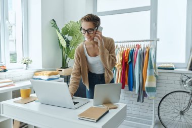 Moda mağazasında çalışırken cep telefonuyla konuşan neşeli genç bir kadın.