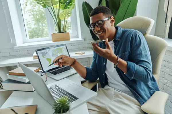 在办公室工作时 快乐的非洲男人一边打电话一边用笔记本电脑 — 图库照片