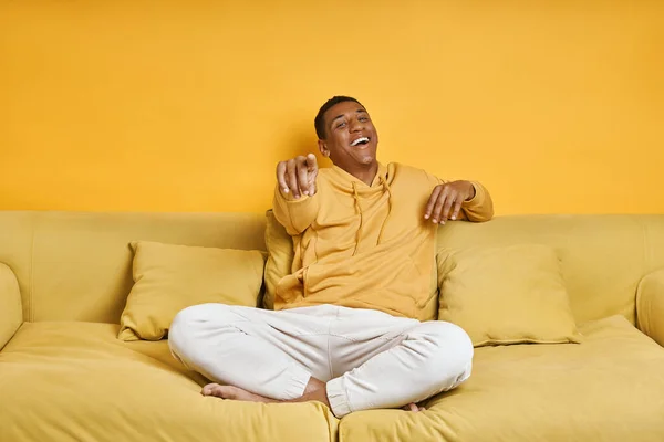 幸せなアフリカ人男性ポインティングカメラながら座ってソファに黄色の背景 — ストック写真