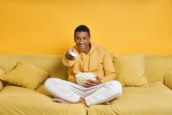 幸せな男を見ますテレビと持っています軽食しながらソファの上でリラックス黄色の背景 — ストック写真