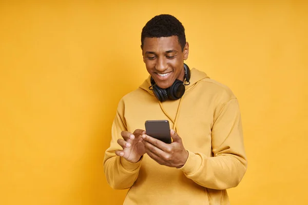 一个戴耳机的英俊的非洲男人站在黄色一边用智能手机 — 图库照片