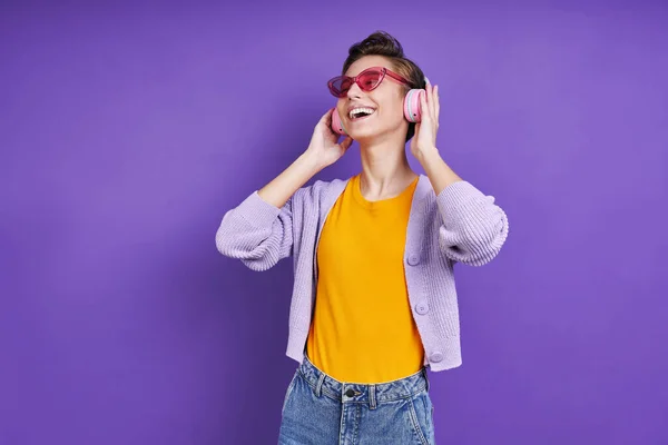 ヘッドフォンの陽気な若い女性は紫の背景を背景に立ちながら音楽を楽しむ — ストック写真