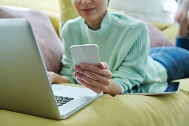 Evdeki kanepede uzanmış akıllı telefon kullanan genç bir kadının yakın çekimi.