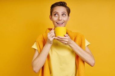Sarı arka planda dururken elinde sıcak içecekle fincanı tutan mutlu genç kadın.
