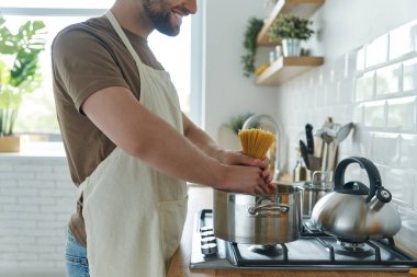 Close-up of young man placing spaghetti into the pan while standing at the domestic kitchen