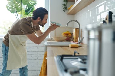Kendine güvenen genç adam bulaşık yıkadıktan sonra mutfak lavabosunun yanında duruyor.