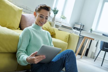 Evde yerde otururken dizüstü bilgisayar kullanan neşeli genç bir kadın.