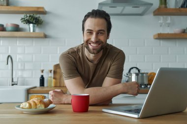 Cheerful man enjoying breakfast and working on laptop while sitting at the kitchen island