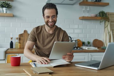 Evdeki mutfak adasında çalışırken teknoloji kullanan neşeli genç adam.