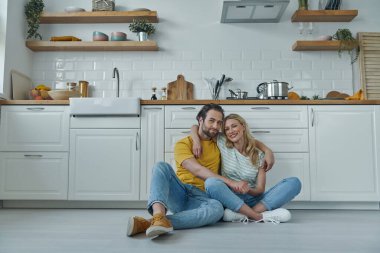 Genç ve güzel bir çift yerde oturup mutfak tezgahına yaslanırken kucaklaşıyorlar.