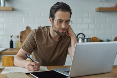 Evdeki mutfak adasında otururken dizüstü bilgisayarda çalışan konsantre genç adam.