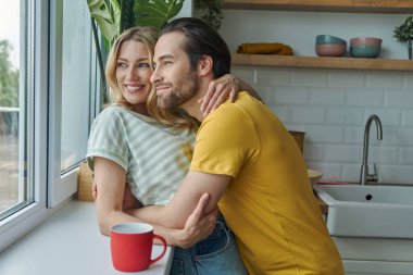 Beautiful young couple looking through a window while standing at the kitchen