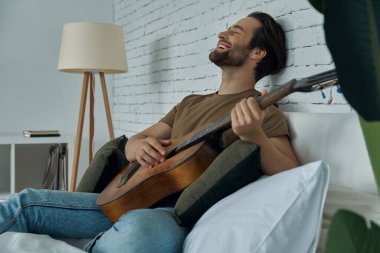 Mutlu genç adam evde yatakta dinlenirken akustik gitar çalıyor.