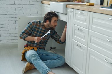 Ev mutfağında lavabo tamir ederken İngiliz anahtarı kullanan kendinden emin bir genç.