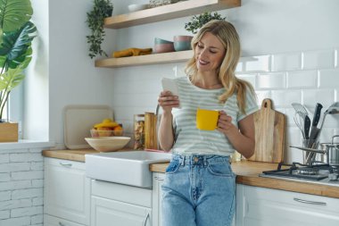 Mutfakta kahve içerken akıllı telefon kullanan neşeli genç bir kadın.