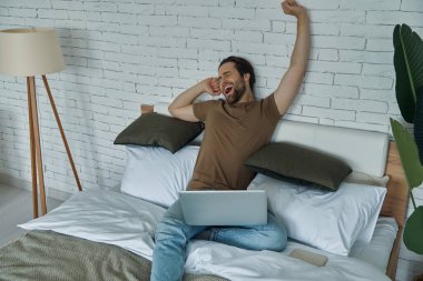 Evde dizüstü bilgisayarla oturup esnemekten sıkılmış genç bir adam.