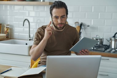 Yoğunlaşmış adam cep telefonuyla konuşuyor ve evde çalışırken teknoloji kullanıyor.