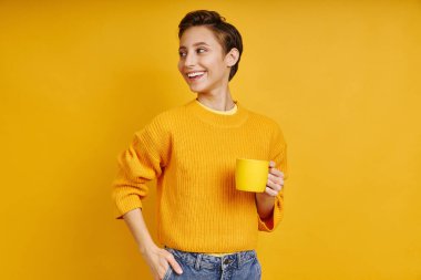 Cheerful young woman holding cup with hot drink while standing against yellow background