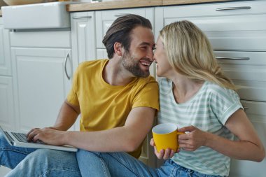 Happy loving couple using laptop while sitting on the floor and leaning at kitchen counter