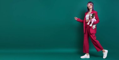 Yeşil arka planda yürürken kucağında küçük bir köpek taşıyan göz kamaştırıcı genç kadın.