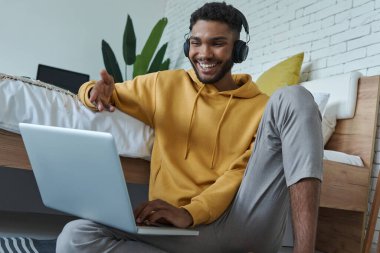 Neşeli Afrikalı adam evde yerde otururken internet konferansı yapıyor ve el kol hareketi yapıyor.