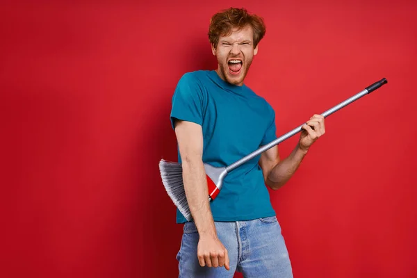 Happy Young Redhead Man Using Broom Guitar While Red Background — Foto Stock