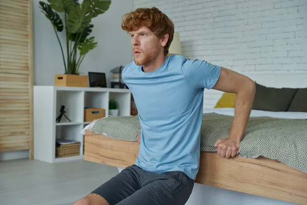 Concentrated Young Redhead Man Doing Push Ups Using Bed Home — Stock fotografie
