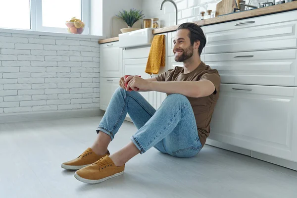 Cheerful Young Man Enjoying Coffee While Sitting Floor Leaning Kitchen — Stockfoto