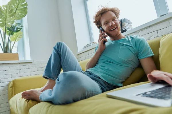 Jistý Zrzka Muž Mluví Telefonu Pomocí Notebooku Zatímco Sedí Gauči — Stock fotografie