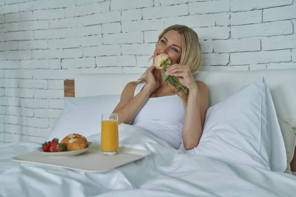 Attractive Young Woman Smelling Flower While Enjoying Breakfast Bed — Stockfoto