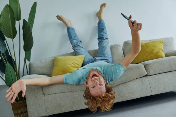 Zabawny Rudzielec Trzyma Smartfon Gestykuluje Relaksując Się Kanapie Domu — Zdjęcie stockowe
