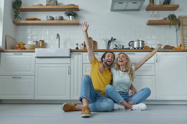 Cheerful Couple Embracing Keeping Arms Raised While Sitting Floor Kitchen — Zdjęcie stockowe