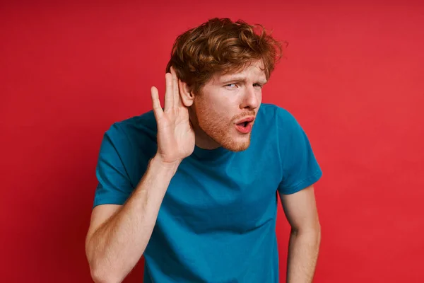 Concentrated Redhead Man Listening While Holding Hand Ear Red Background — Stock Photo, Image