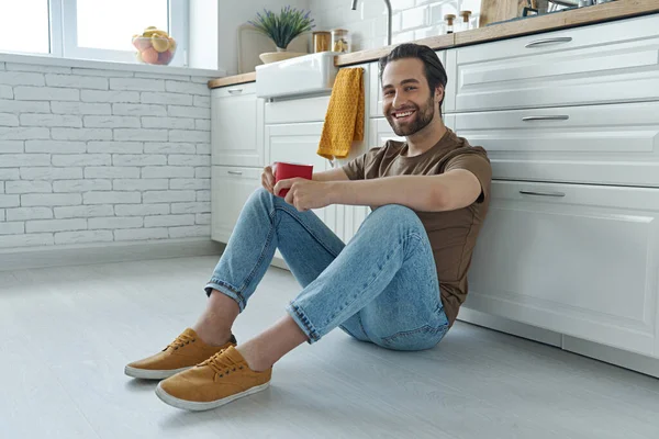 Happy Young Man Enjoying Coffee While Sitting Floor Leaning Kitchen — Fotografia de Stock