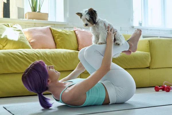 Confident woman in sports clothing exercising with her little dog at home