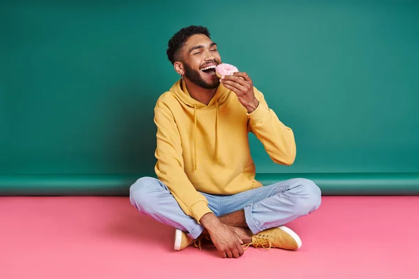 Cheerful African Man Enjoying Doughnut While Sitting Colorful Background — ストック写真