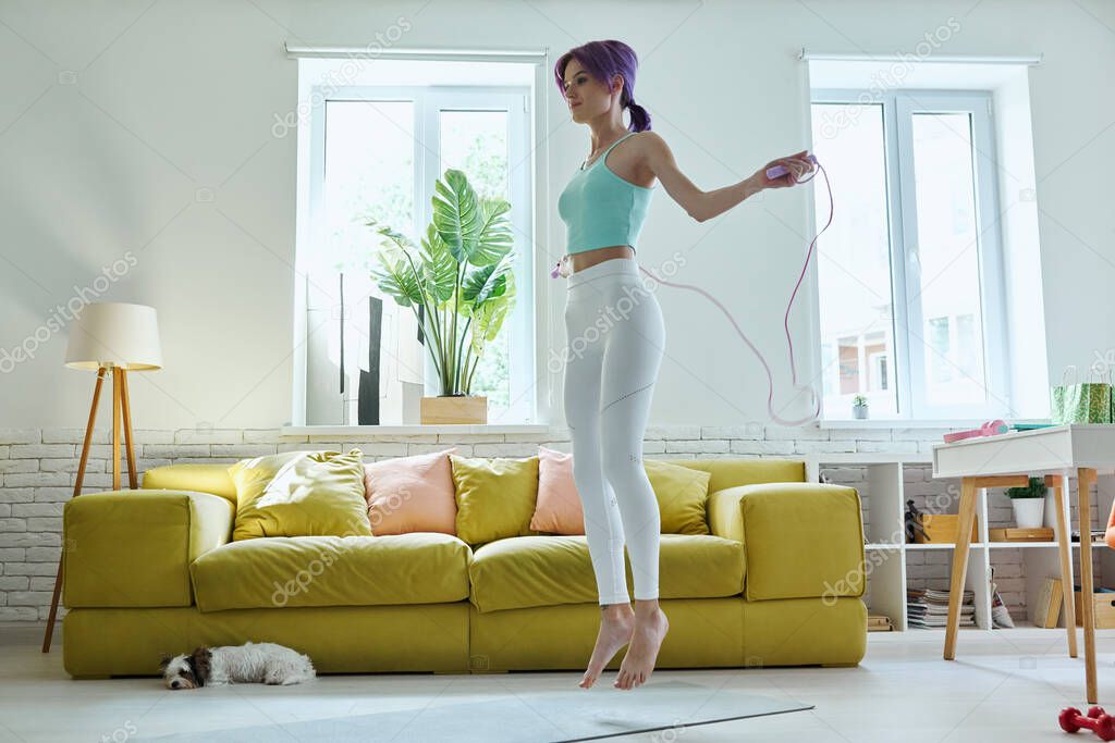 Confident young woman in sports clothing jumping with rope at home