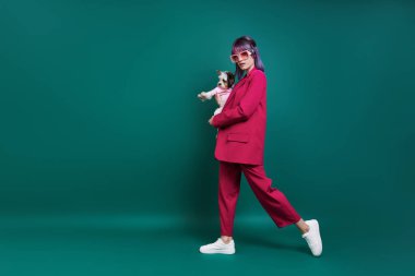 Yeşil arka planda yürürken kucağında küçük bir köpek taşıyan göz kamaştırıcı genç kadın.