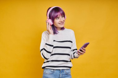 Sarı arka planda dururken akıllı telefon kullanan, kulaklıklı neşeli genç bir kadın.