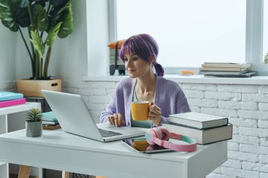 Mor saçlı, kendine güveni tam genç bir kadın çalıştığı yerde bilgisayarla çalışıyor.
