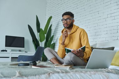 Genç Afrikalı adam not defterini tutuyor ve evde yatakta otururken düşünceli görünüyor.