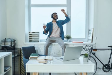 Ofisteki pencere eşiğinde otururken akıllı telefon kullanan kendine güvenen bir Afrikalı.