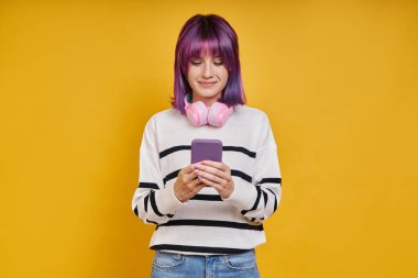 Kulaklıklı, sarı arka planda akıllı telefon kullanan kendine güvenen genç bir kadın.