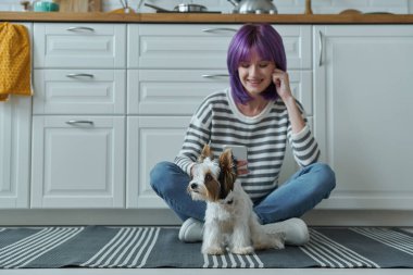 Cheerful woman using smart phone while spending time with her dog at the kitchen